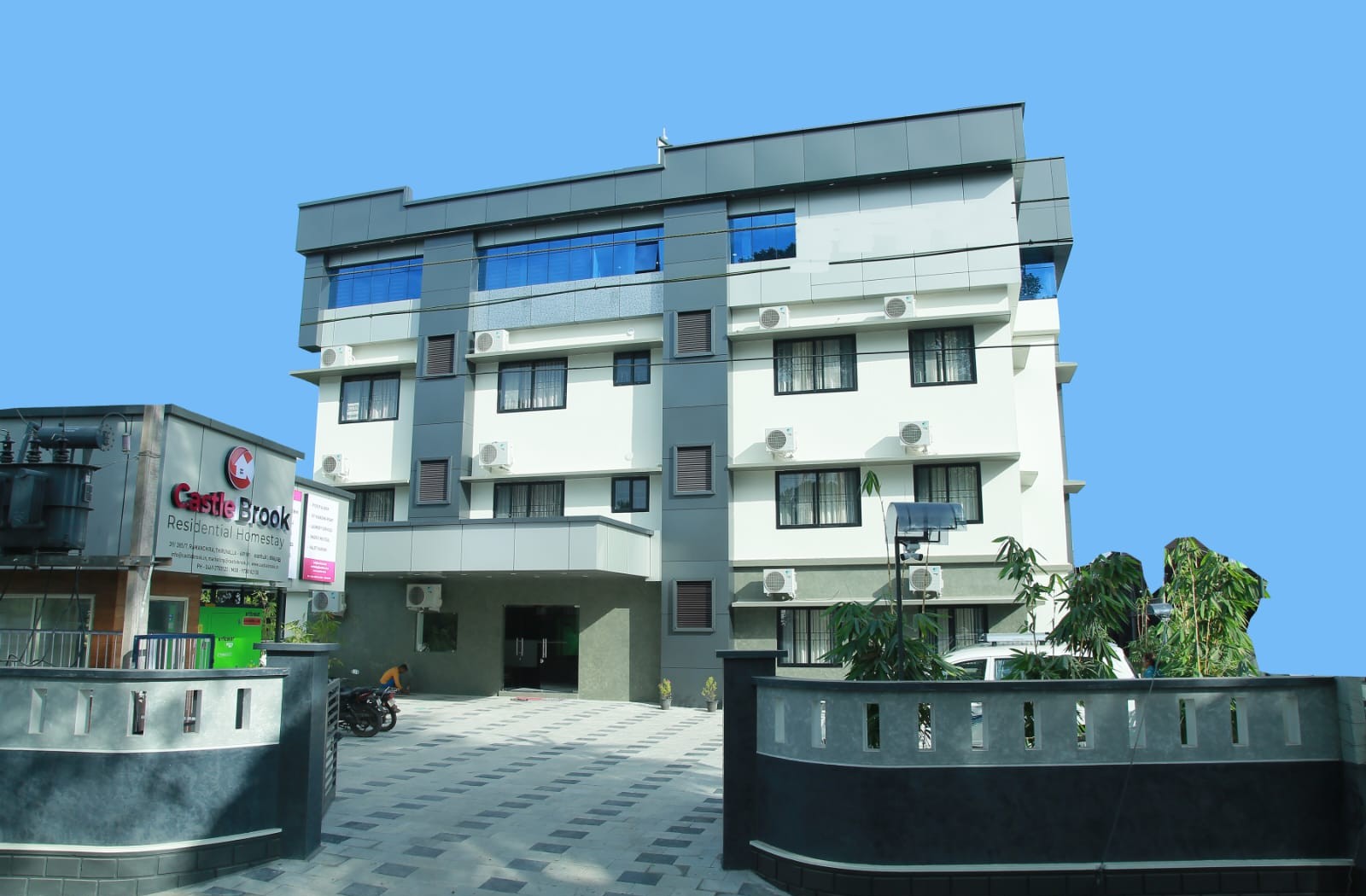 xterior view of a modern multi-storey building labeled Castle Brook Residential Homestay, with a tiled driveway and surrounding greenery.